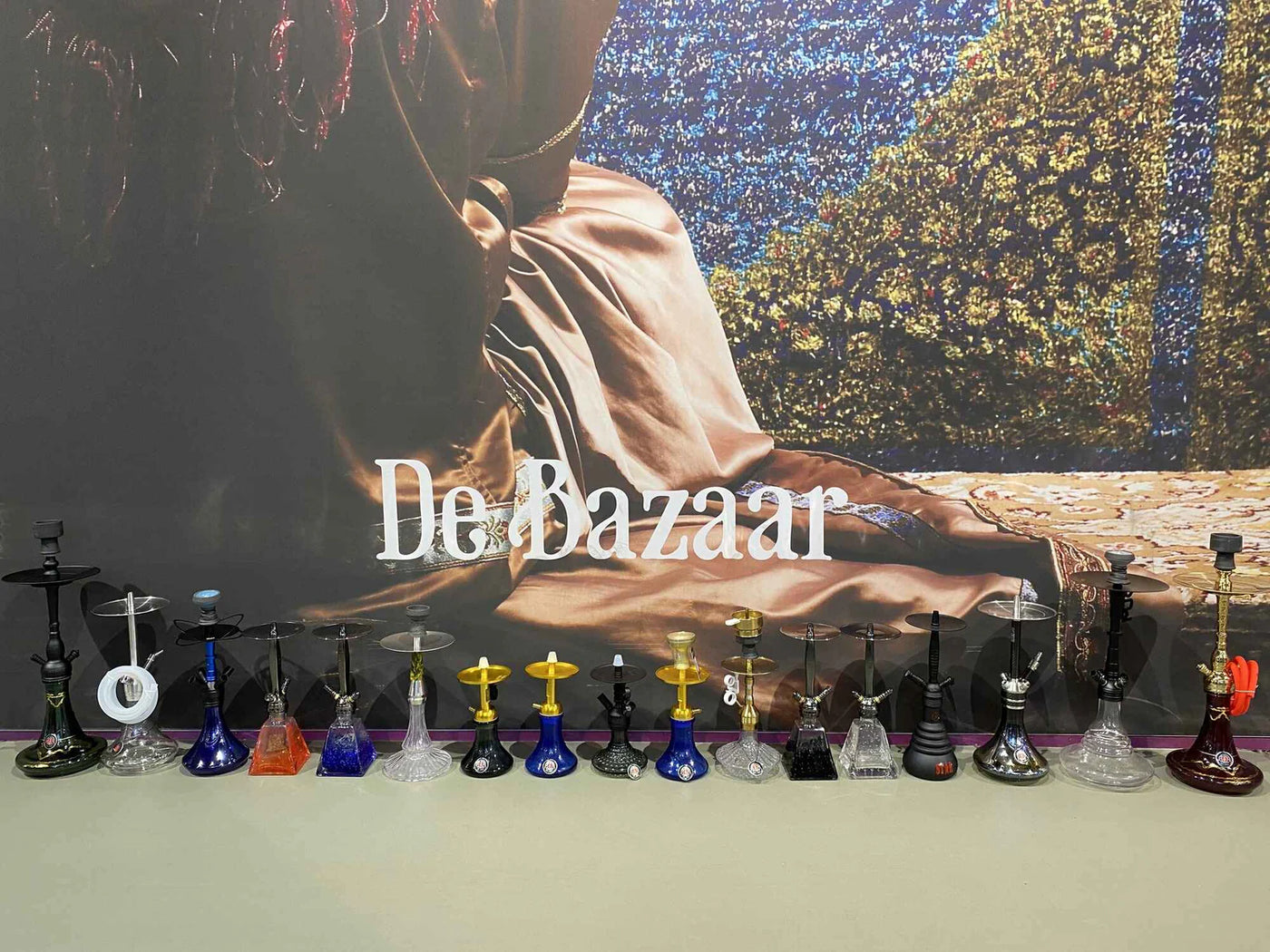Rij glazen waterpijpen op de toonbank van onze shisha winkel op de Bazaar in Beverwijk, voor een grote wandfoto met de tekst De Bazaar.