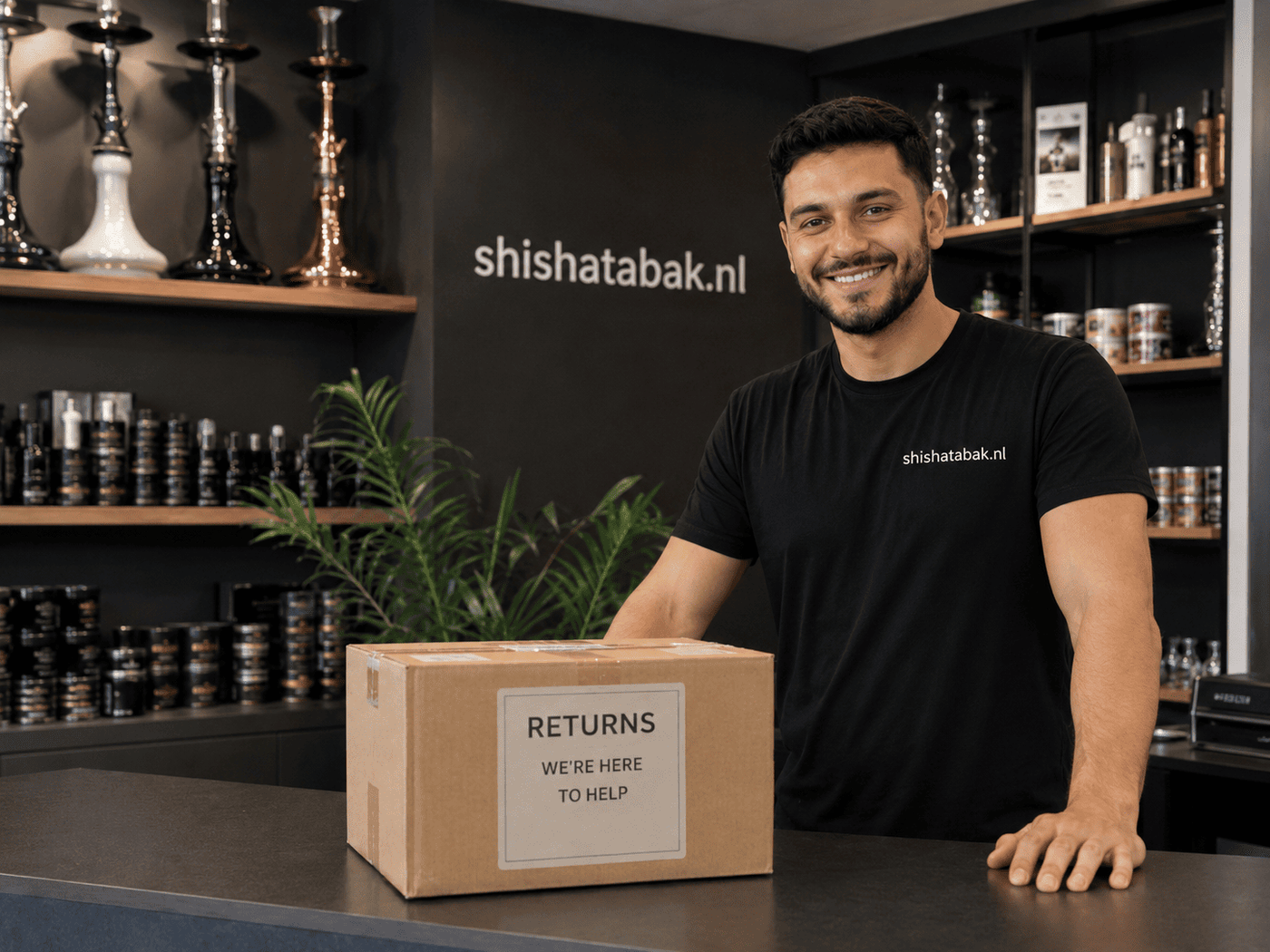 Lachende ShishaTabak medewerker achter de toonbank met een retourdoos, voor de wand met waterpijpen en shisha tabak. Klachten en retours van je bestelling handelen we gratis af.